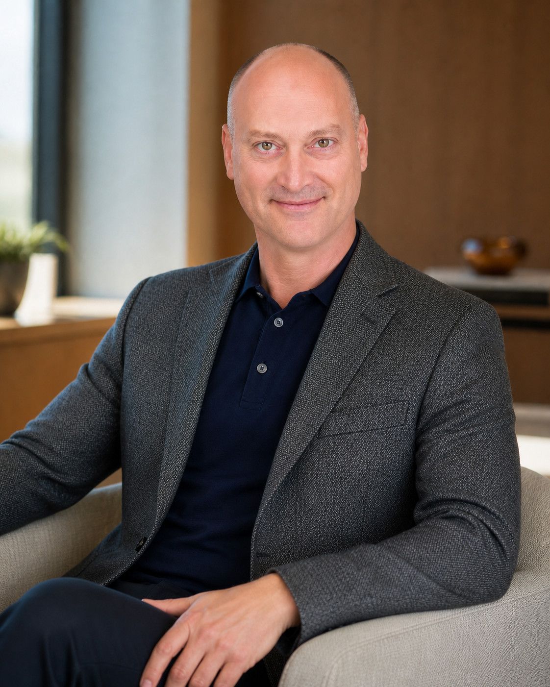 Peter DeCaprio seated in a pale armchair in a wood-panelled lounge, wearing a charcoal tweed jacket over a navy polo shirt
