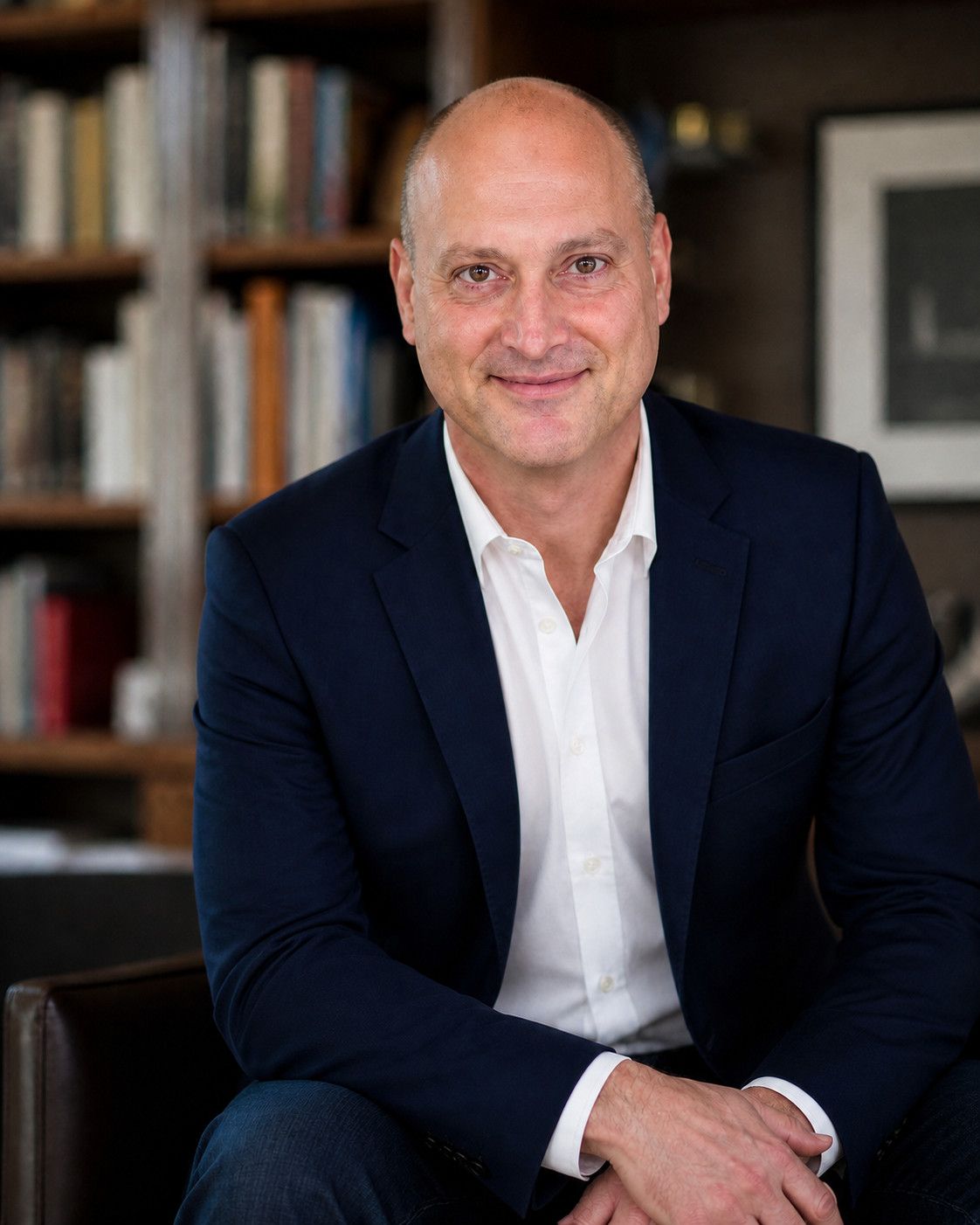 Peter DeCaprio seated in a leather armchair in front of a wall of bookshelves, in a navy jacket and open white shirt, smiling at the camera